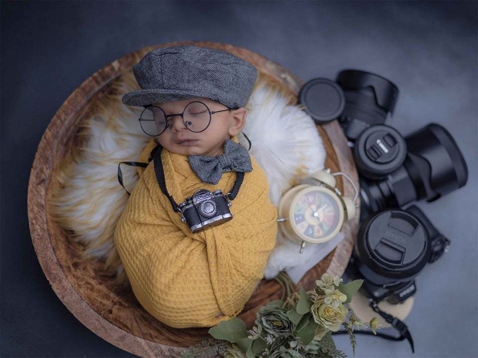 Cute newborn baby sleeping in a wooden bowl with a mini camera and DSLR lenses in a photographer-themed setup