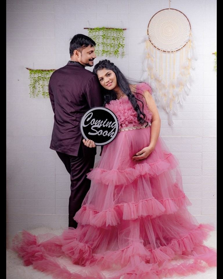 Pregnant woman in a pink gown leaning against her husband with a “Coming Soon” sign in a romantic studio portrait